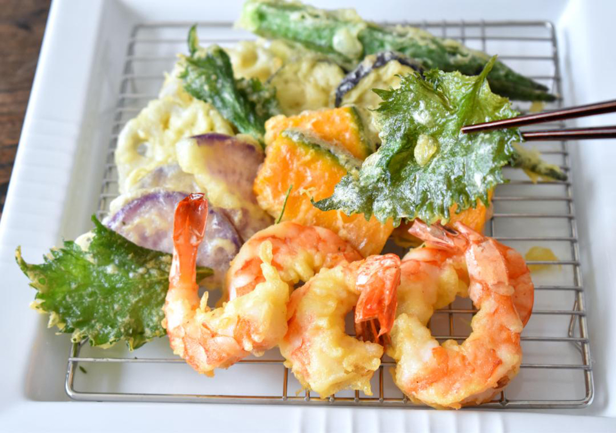 米粉で作ったえびやなす、かぼちゃなどの天ぷらがお皿に盛られ、大葉の天ぷらを箸でつまんでいる様子